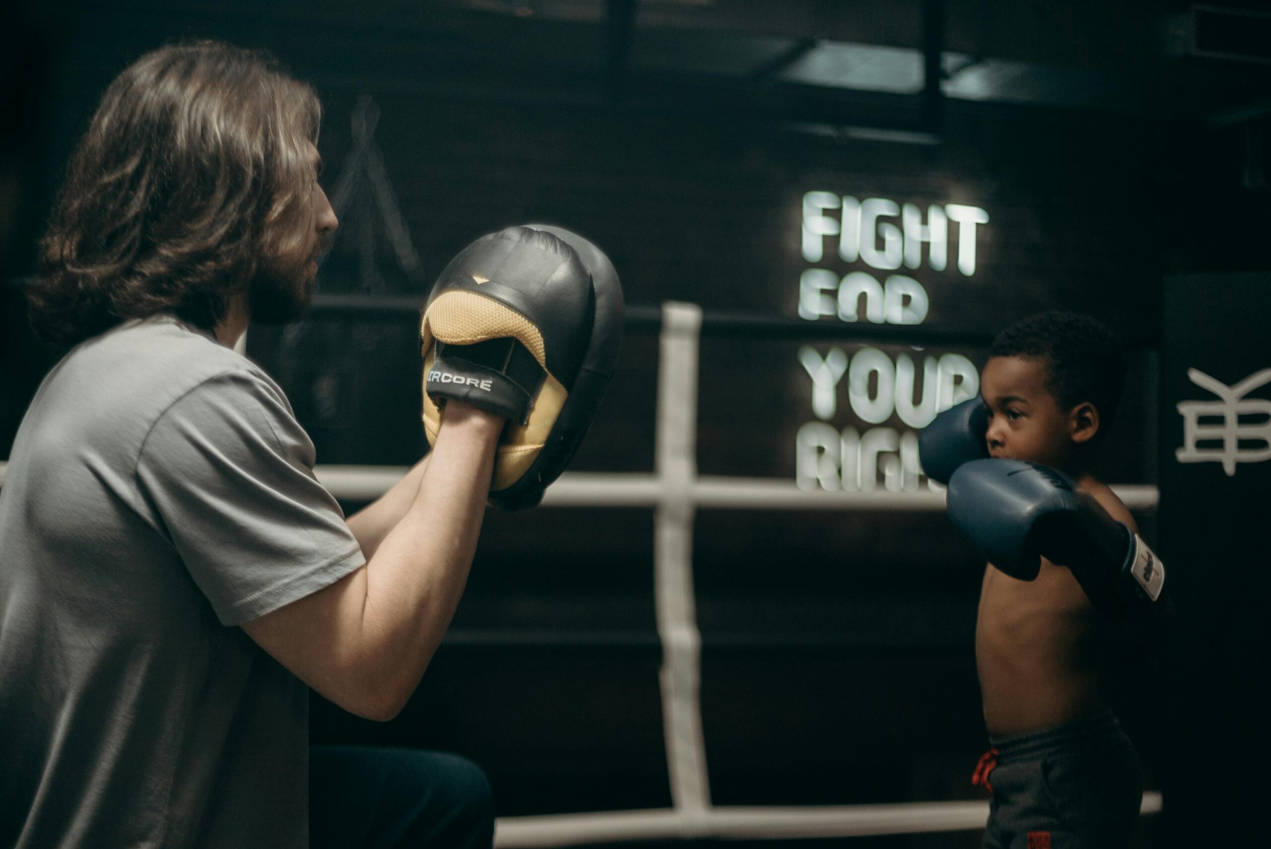 A young boy in boxing gloves trains with his coach in a gym, sign reads 'Fight For Your Right'.